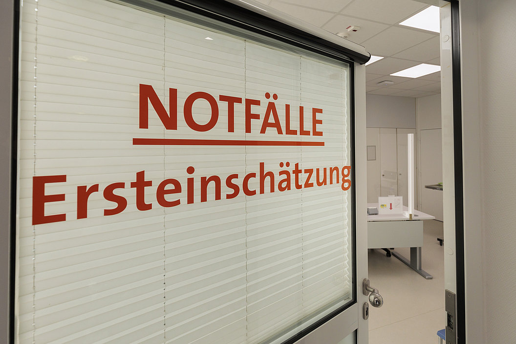 Tür mit Sichtschutzfolie, darauf in roter Schrift 'NOTFÄLLE Ersteinschätzung'. Im Hintergrund ein Büro mit Schreibtisch und Stuhl.
