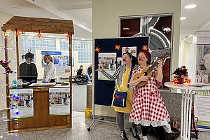 Zwei Frauen stehen in einem Innenraum, eine spielt Ukulele, die andere zeigt auf ein Plakat an einer Stellwand, im Hintergrund ein Stand mit Informationsmaterial und Lichterketten