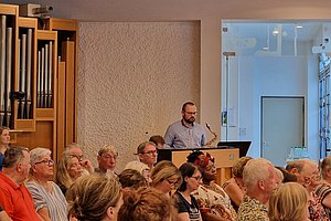 Person spielt Saxophon an einem Rednerpult vor einem Publikum in einem Raum mit Orgelpfeifen an der Wand