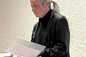 Eine Person mit langen, grauen Haaren, die zu einem Pferdeschwanz gebunden sind, steht an einem Rednerpult und hält ein Dokument in der Hand. Die Person trägt eine schwarze Jacke und ein schwarzes Hemd.