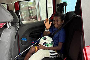 Ein Kind sitzt auf dem Rücksitz eines roten Fahrzeugs, hält einen Fußball und winkt. Das Kind trägt ein blaues T-Shirt und hat einen Gipsverband am Bein.