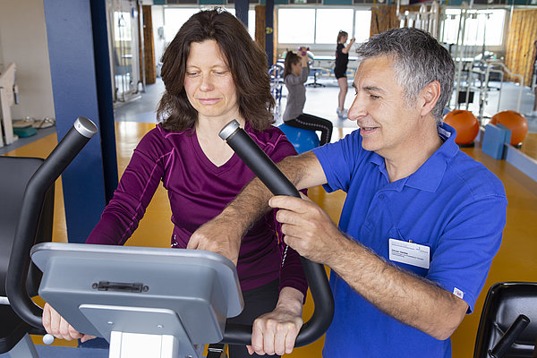 Frau auf einem Ergometer mit männlichem Trainer, der sie anleitet, im Fitnessraum mit weiteren Personen im Hintergrund