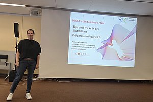 Person steht vor einer Leinwand mit einer Präsentation zu 'Tips und Tricks in der Blutstillung' und 'Präparate im Vergleich' von Rebecca Schweizer.
