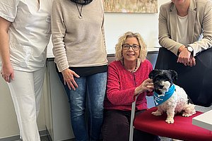 Vier Personen in einem Raum mit einem roten Stuhl, auf dem ein kleiner schwarz-weißer Hund mit blauem Halstuch sitzt. Im Hintergrund ein Bild mit roten Mohnblumen.