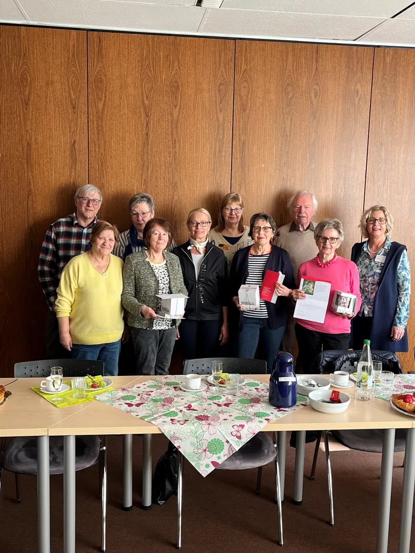 Gruppe von elf Personen steht hinter einem Tisch mit Kaffeetassen, Thermoskanne und gedeckter Tischdecke in einem Raum mit Holzpaneelen