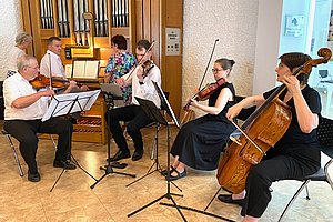 Fünf Musiker spielen in einem Raum mit Orgel im Hintergrund, darunter zwei Geiger, eine Cellistin und eine Flötistin, alle sitzen auf Stühlen und lesen von Notenständern.