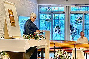 Person in schwarzem Gewand steht an einem mit weißem Tuch bedeckten Altar mit Blumen, im Hintergrund bunte Kirchenfenster und eine weitere Person in weißem Gewand auf einer Bank.