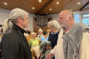 Zwei Personen im Gespräch in einem Raum mit Holzbänken und einer Orgel im Hintergrund. Eine Person trägt einen grauen Pullover und eine andere hat graue Haare.
