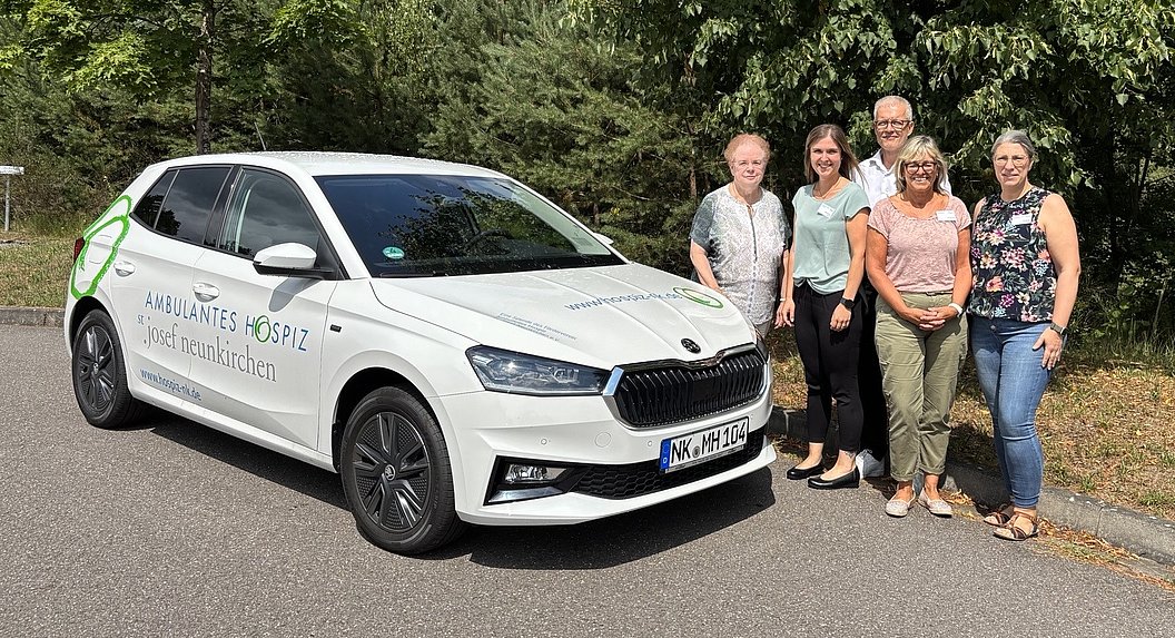 Weißer Dienstwagen mit der Aufschrift »Ambulantes Hospiz St. Josef Neunkirchen« steht auf einer Straße neben einer Gruppe von fünf Personen vor grünem Hintergrund