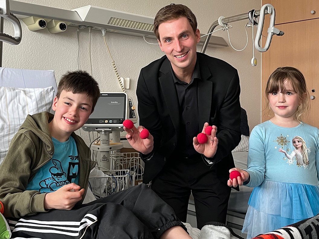 Drei Personen in einem Krankenzimmer, eine sitzend auf einem Bett, die andere zwei stehen daneben und halten jeweils mehrere rote Bälle in den Händen