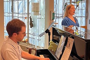 Zwei Personen in einem Raum mit großen Fenstern, eine spielt Klavier, die andere steht daneben