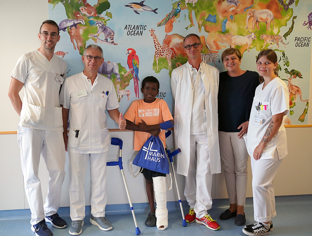 Gruppe von fünf Personen in weißen Kitteln, darunter ein Junge mit Gipsverband am Bein, der auf einem Gehstock steht. Im Hintergrund eine Weltkarte mit Tieren.