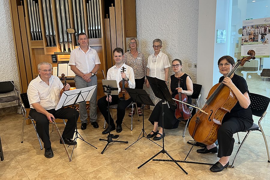 Sechs Personen mit Streichinstrumenten und Notenständern sitzen und stehen vor einer großen Orgel in einem Raum