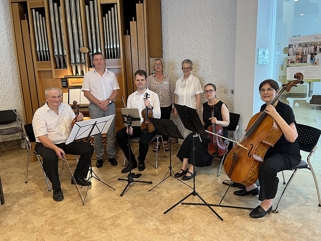 Sechs Personen mit Streichinstrumenten und Notenständern sitzen und stehen vor einer großen Orgel in einem Raum