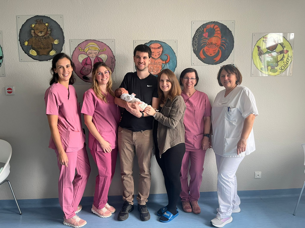 Gruppe von Personen in medizinischer Kleidung und Zivilkleidung, eine Person hält ein Baby, bunte Tierbilder an der Wand im Hintergrund.
