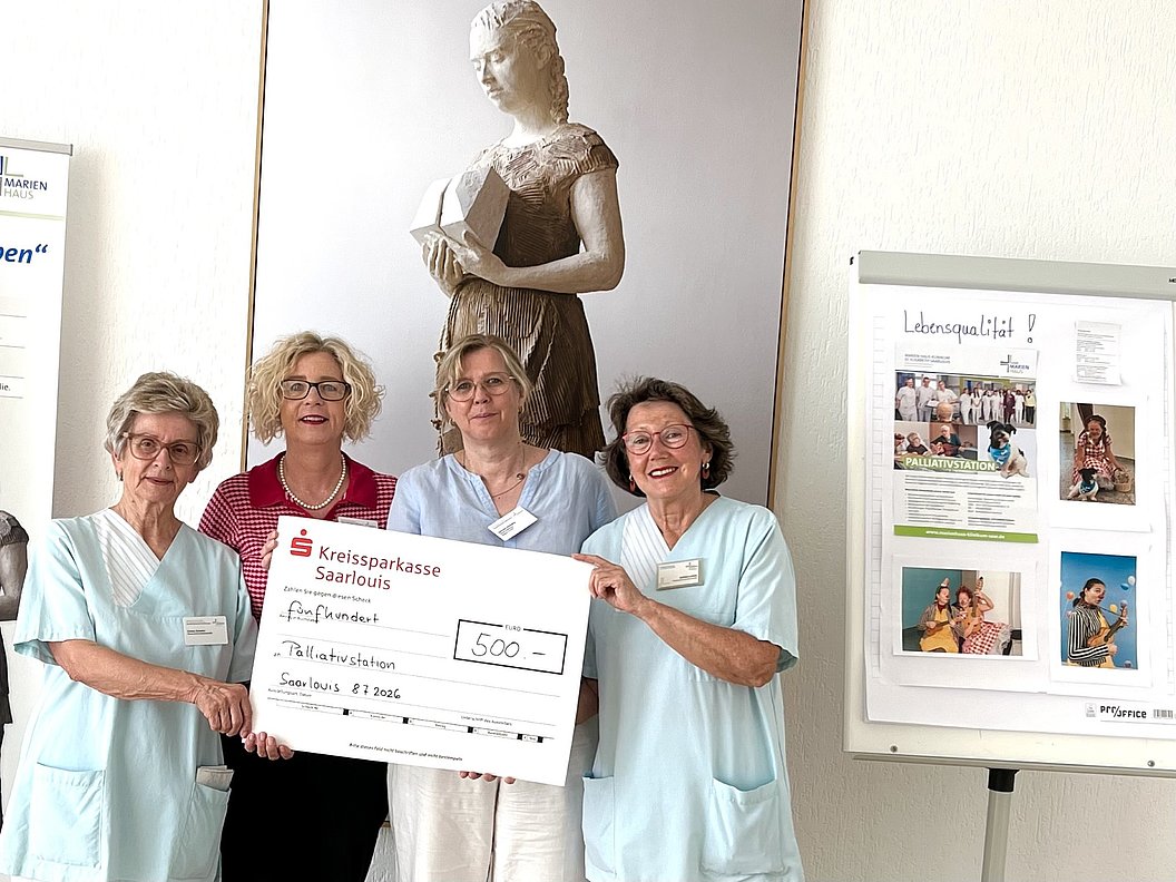 Vier Frauen in medizinischer Arbeitskleidung und eine Frau in rotem Oberteil halten gemeinsam einen großen symbolischen Scheck vor einer Statue und einem Informationsplakat