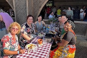 Fünf Personen sitzen an einem Tisch mit rot-weiß kariertem Tuch im Freien unter einer Holzkonstruktion, essen und unterhalten sich.