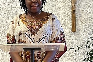 Frau in traditionellem afrikanischem Kleid steht hinter einem Rednerpult in einer Kirche, im Hintergrund ein Kruzifix an der Wand