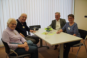 Vier Personen sitzen an einem rechteckigen Tisch in einem Raum mit vertikalen Jalousien und haben Getränke vor sich stehen