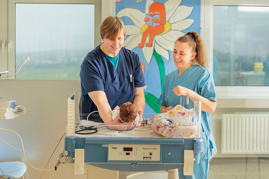 Zwei medizinische Fachkräfte in blauer Arbeitskleidung betreuen ein Neugeborenes auf einem Inkubator im Krankenhauszimmer mit bunter Wandmalerei