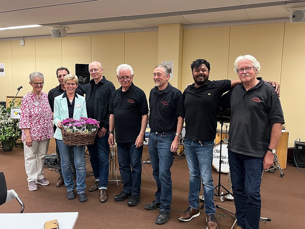 Sieben Personen stehen nebeneinander in einem Raum mit Mikrofonständern und Musikinstrumenten im Hintergrund, eine Person hält einen Korb mit Blumen.