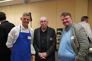 Drei Männer in einem Raum, einer trägt eine blaue Schürze, die anderen zwei tragen Jackett und Pullover, im Hintergrund weitere Personen und ein Regal mit Getränkekisten