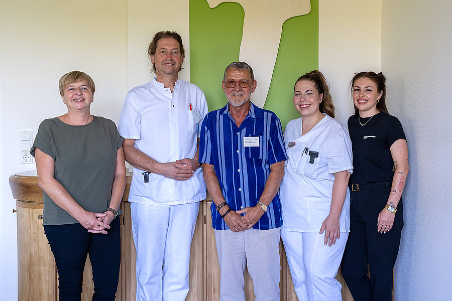 Fünf Personen stehen nebeneinander vor einer hellen Wand mit grünem Bild, zwei tragen weiße medizinische Kleidung, die anderen legere Kleidung
