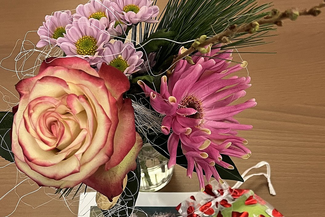 Blumenstrauß mit rosa Rose und pinken Chrysanthemen neben einer Karte mit Text und einem kleinen Geschenk mit Herzmuster auf Holztisch.