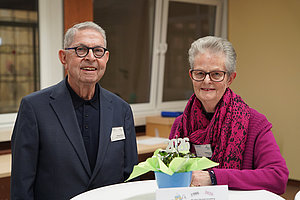 Älterer Mann und ältere Frau mit grauen Haaren und rotem Schal stehen an einem Stehtisch mit Blumenarrangement und Namensschild