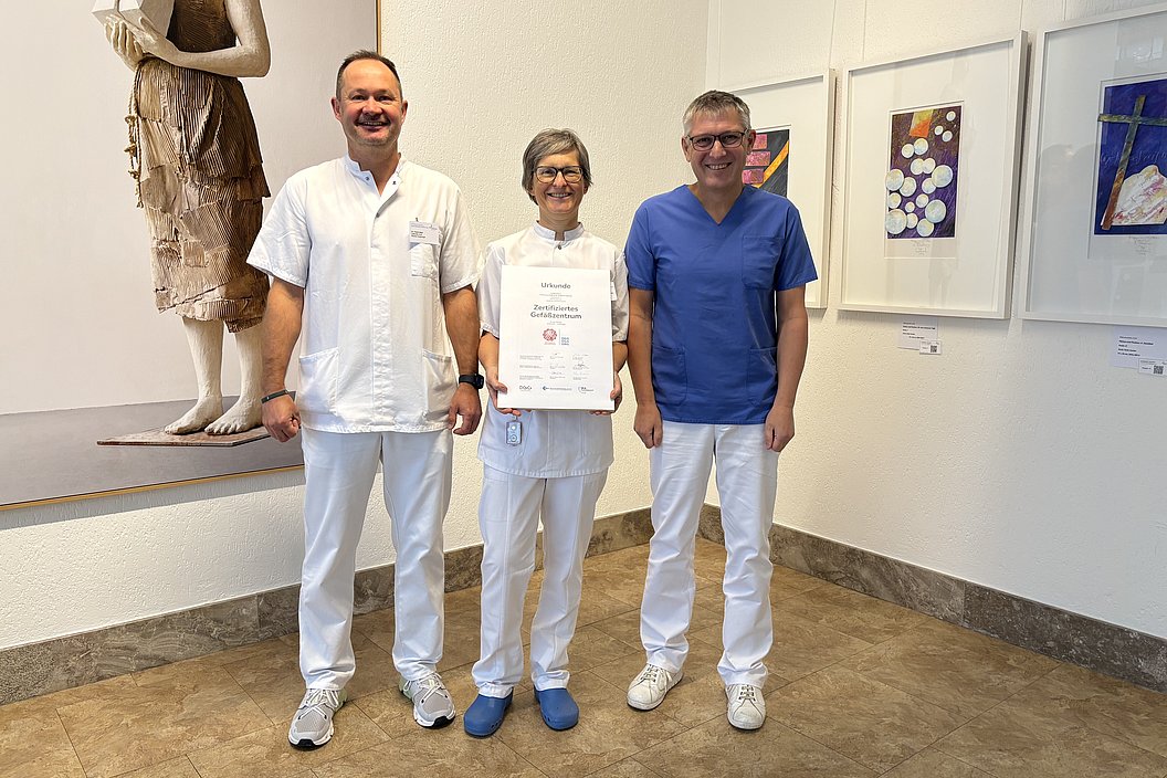 Drei Personen in medizinischer Kleidung stehen in einem Raum mit Kunstwerken an der Wand. Frau in der Mitte hält ein Zertifikat in den Händen.