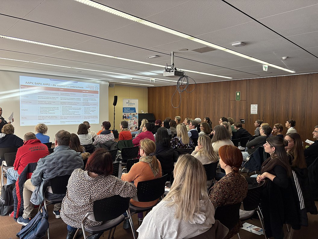Gruppe von Menschen sitzt in einem Seminarraum und schaut auf eine Projektionsfläche mit Text und Tabellen