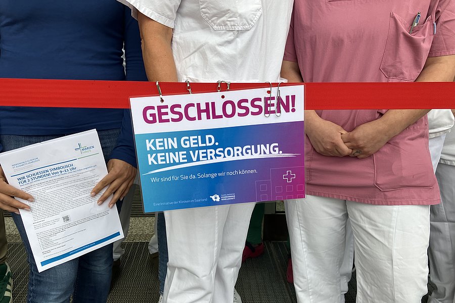 Drei Personen in medizinischer Kleidung stehen hinter einem roten Absperrband mit einem Schild 'GESCHLOSSEN! KEIN GELD. KEINE VERSORGUNG.'