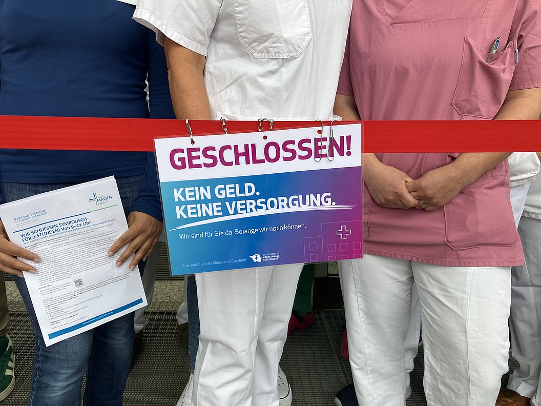 Drei Personen in medizinischer Kleidung stehen hinter einem roten Absperrband mit einem Schild 'GESCHLOSSEN! KEIN GELD. KEINE VERSORGUNG.'