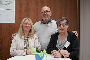 Drei Personen stehen an einem runden Tisch mit einer blauen Vase und einer Pflanze darauf vor einer Holzvertäfelung mit einem Aushang an der Wand