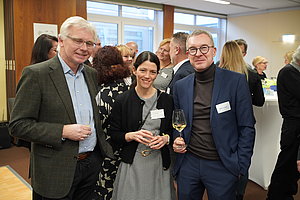 Gruppe von Menschen in Businesskleidung bei einer Veranstaltung mit Getränken in der Hand in einem hellen Raum mit großen Fenstern