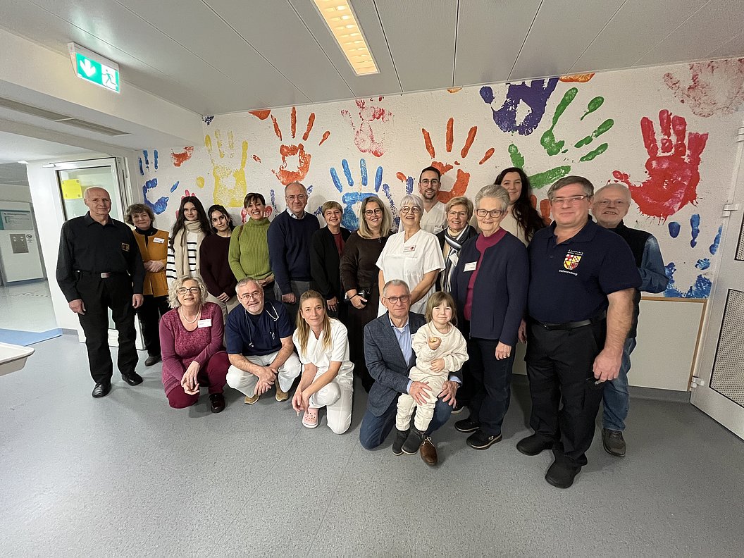 Gruppe von Erwachsenen und Kindern steht vor einer Wand mit bunten Handabdrücken in verschiedenen Größen.