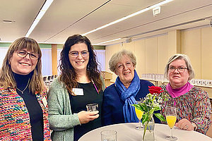 Vier Frauen stehen um einen hohen, runden Tisch mit einer Vase, die Blumen enthält, und mehreren Gläsern mit Getränken.