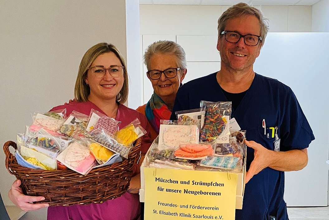 Zwei Personen in medizinischer Kleidung halten einen Korb und eine Kiste mit kleinen, verpackten Babymützen und Strümpfen, Aufschrift auf der Kiste: 'Mützchen und Strümpfchen für unsere Neugeborenen'.