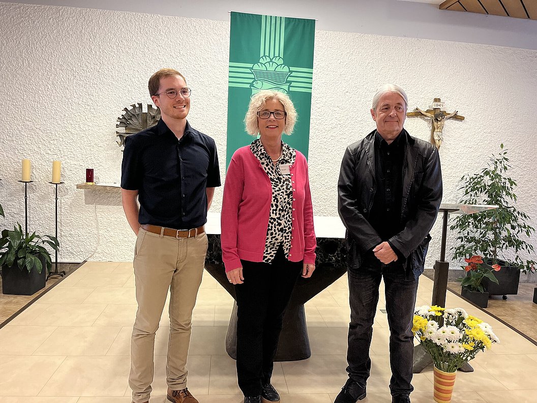 Drei Personen stehen vor einem Altar in einem Raum. Links ein Mann in schwarzem Hemd und hellen Hosen, in der Mitte eine Frau in rosa Cardigan mit gemusterter Bluse, rechts ein Mann in schwarzer Jacke und Hemd. Im Hintergrund sind Pflanzen und Kerzen sichtbar.
