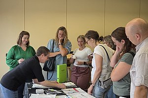Mehrere Personen stehen um einen Tisch mit Papieren und Ordnern, eine Person zeigt auf Dokumente auf dem Tisch.