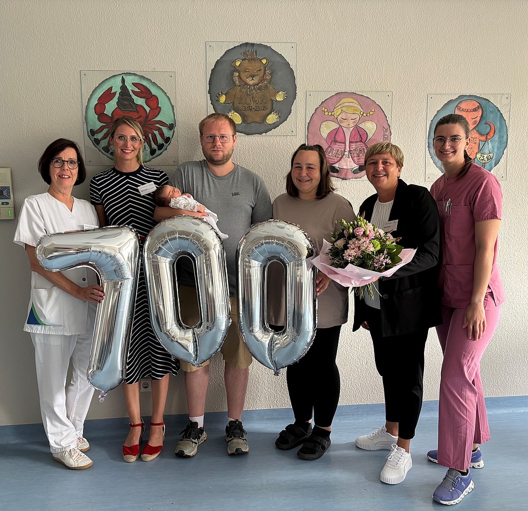 Sechs Personen stehen in einem Raum, drei halten silberne Luftballons, die die Zahl 700 bilden, eine Person hält einen Blumenstrauß, im Hintergrund sind vier Tier- und Engelbilder an der Wand