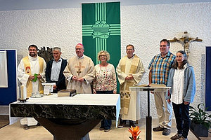 Sieben Personen stehen hinter einem Altar in einer Kirche, darunter drei Geistliche in liturgischer Kleidung, ein grünes Wandtuch mit einem Kreuz und einer Taube, ein Kruzifix an der Wand rechts.
