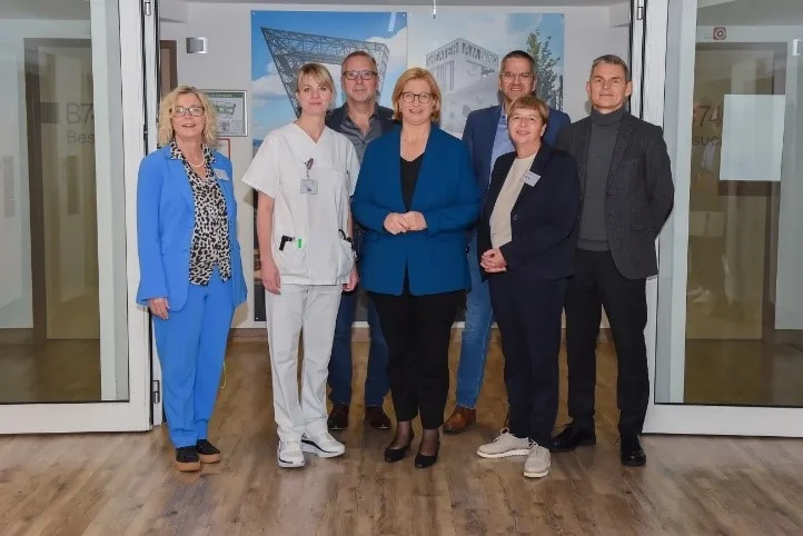 Sieben Personen stehen in einem Flur vor zwei großen Architekturzeichnungen an der Wand, darunter eine Frau in blauem Anzug und eine Person in weißer medizinischer Kleidung.