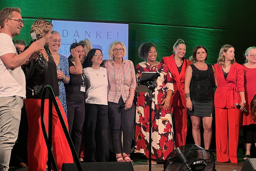 Gruppe von Menschen auf einer Bühne, einige tragen rote Kleidung, andere in verschiedenen Outfits. Im Hintergrund ein Schild mit 'DANKE!'. Mikrofonständer im Vordergrund.