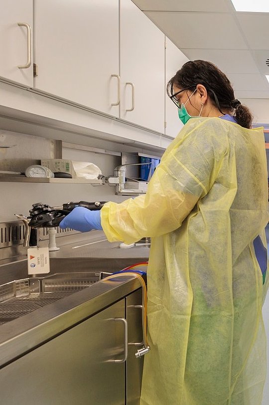 Person in gelbem Schutzkittel, Maske und Handschuhen arbeitet in einer Laborumgebung