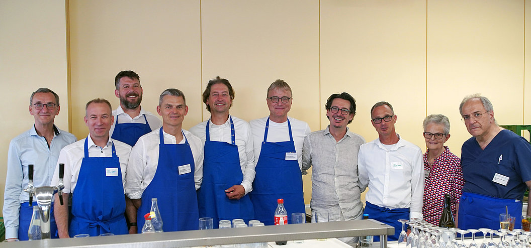 Gruppe von elf Personen, die in einer Reihe stehen, viele tragen blaue Schürzen, vor einem hellen Hintergrund