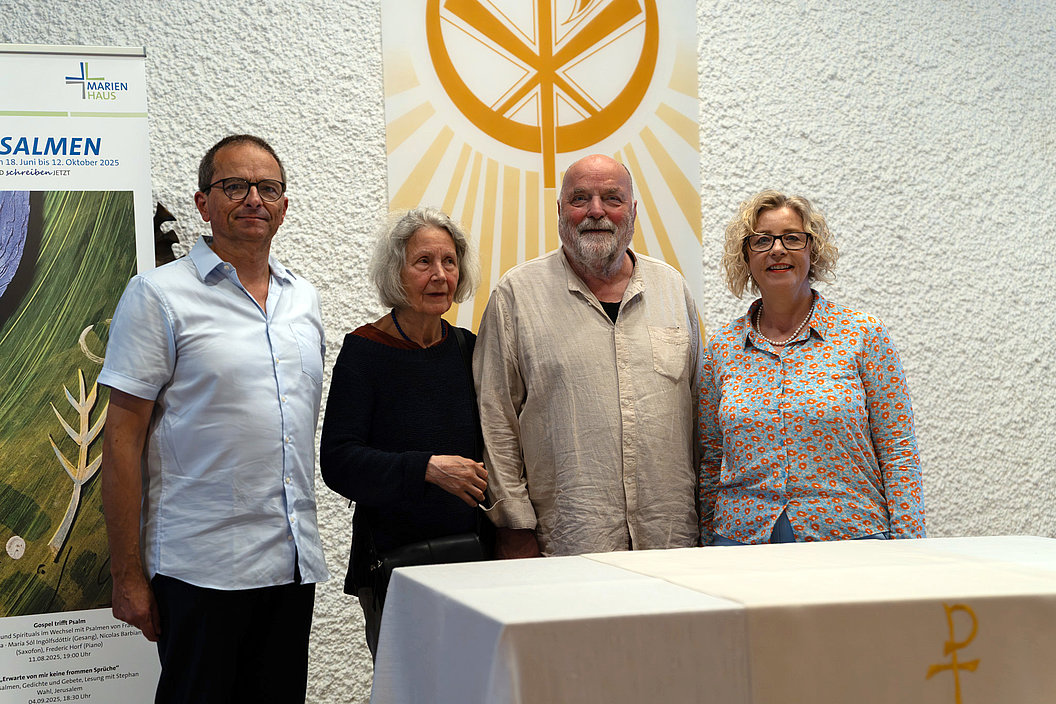 Vier Personen stehen nebeneinander vor einer Wand mit einem gelben christlichen Symbol und einem weißen Altar mit einem goldenen Chi-Rho-Zeichen