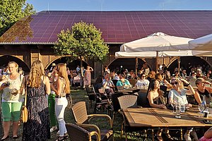 Menschen sitzen und stehen bei einer Gartenparty vor einem Gebäude mit Solardach, Tische mit Sonnenschirmen