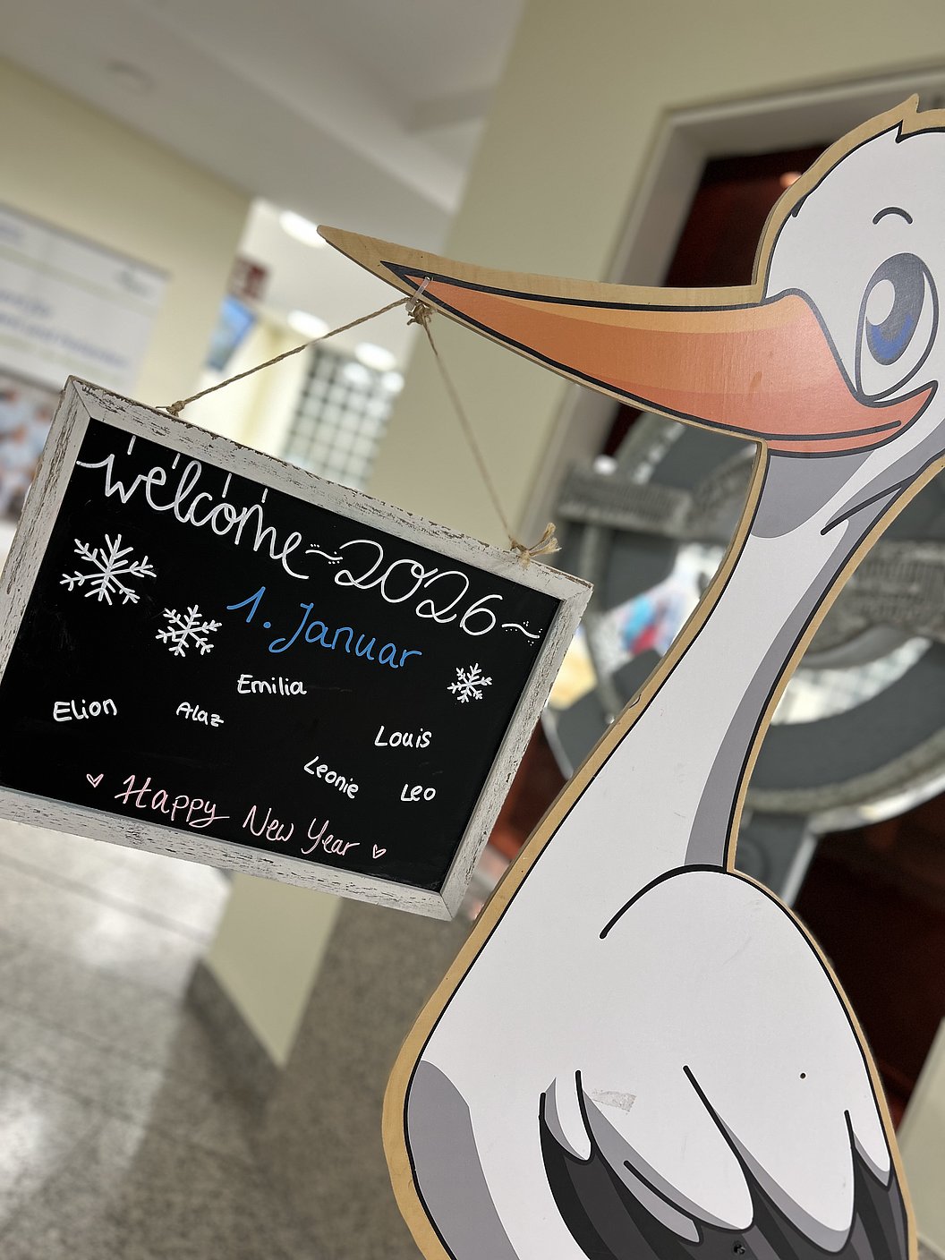 Große Pappfigur eines Storchs hält eine kleine Tafel mit der handgeschriebenen Aufschrift 'welcome 2026 - 1. Januar' und mehreren Namen sowie 'Happy New Year'.