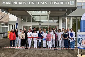 Gruppe von Menschen in medizinischer Kleidung steht vor dem Eingang des Marienhaus Klinikums St. Elisabeth hinter einem roten Absperrband